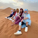 A group of people sitting on a sand dune in the Moroccan desert, wearing traditional clothing and smiling.