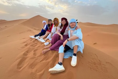 A group of people sitting on a sand dune in the Moroccan desert, wearing traditional clothing and smiling.
