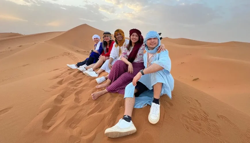 A group of people sitting on a sand dune in the Moroccan desert, wearing traditional clothing and smiling.