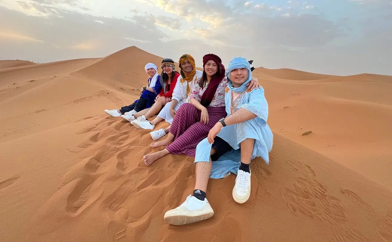 A group of people sitting on a sand dune in the Moroccan desert, wearing traditional clothing and smiling.