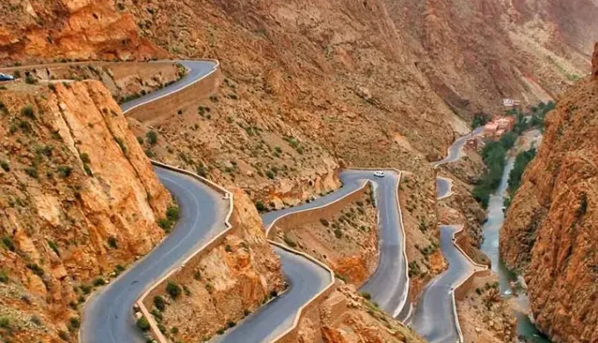 A winding road with sharp switchbacks navigates a rocky canyon, with a river running alongside and sparse vegetation.