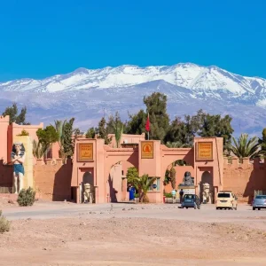 The entrance to a film studio complex in Ouarzazate, Morocco, with statues and signs, set against a backdrop of snow-capped mountains. Keywords: Ouarzazate Film Studios, Morocco, entrance, statues, mountains.