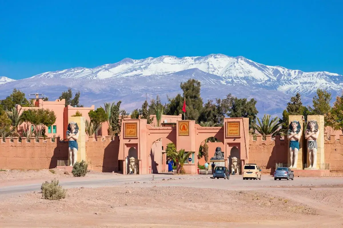 The entrance to a film studio complex in Ouarzazate, Morocco, with statues and signs, set against a backdrop of snow-capped mountains. Keywords: Ouarzazate Film Studios, Morocco, entrance, statues, mountains.