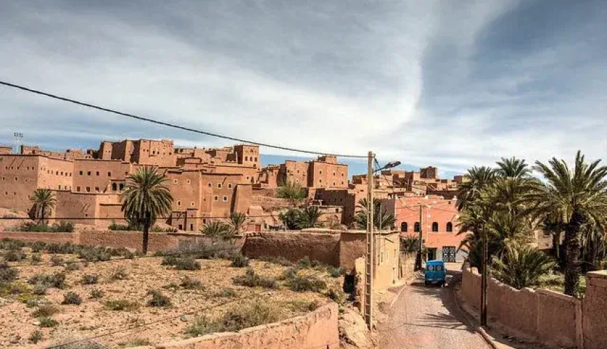 Traditional architecture in Ouarzazate, Morocco, featuring mud-brick buildings, palm trees, and a narrow road. The image highlights the cultural heritage and picturesque landscape of the region.