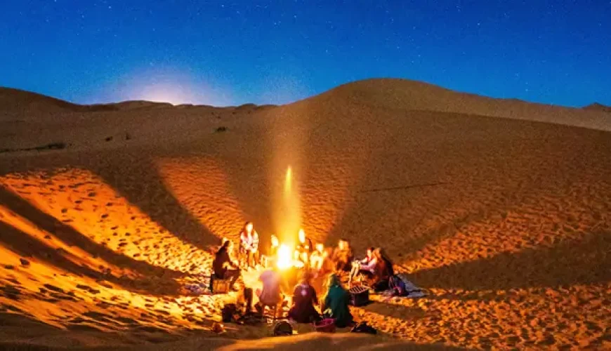 A group of people gather around a campfire in a desert at night, surrounded by sand dunes under a clear, starry sky