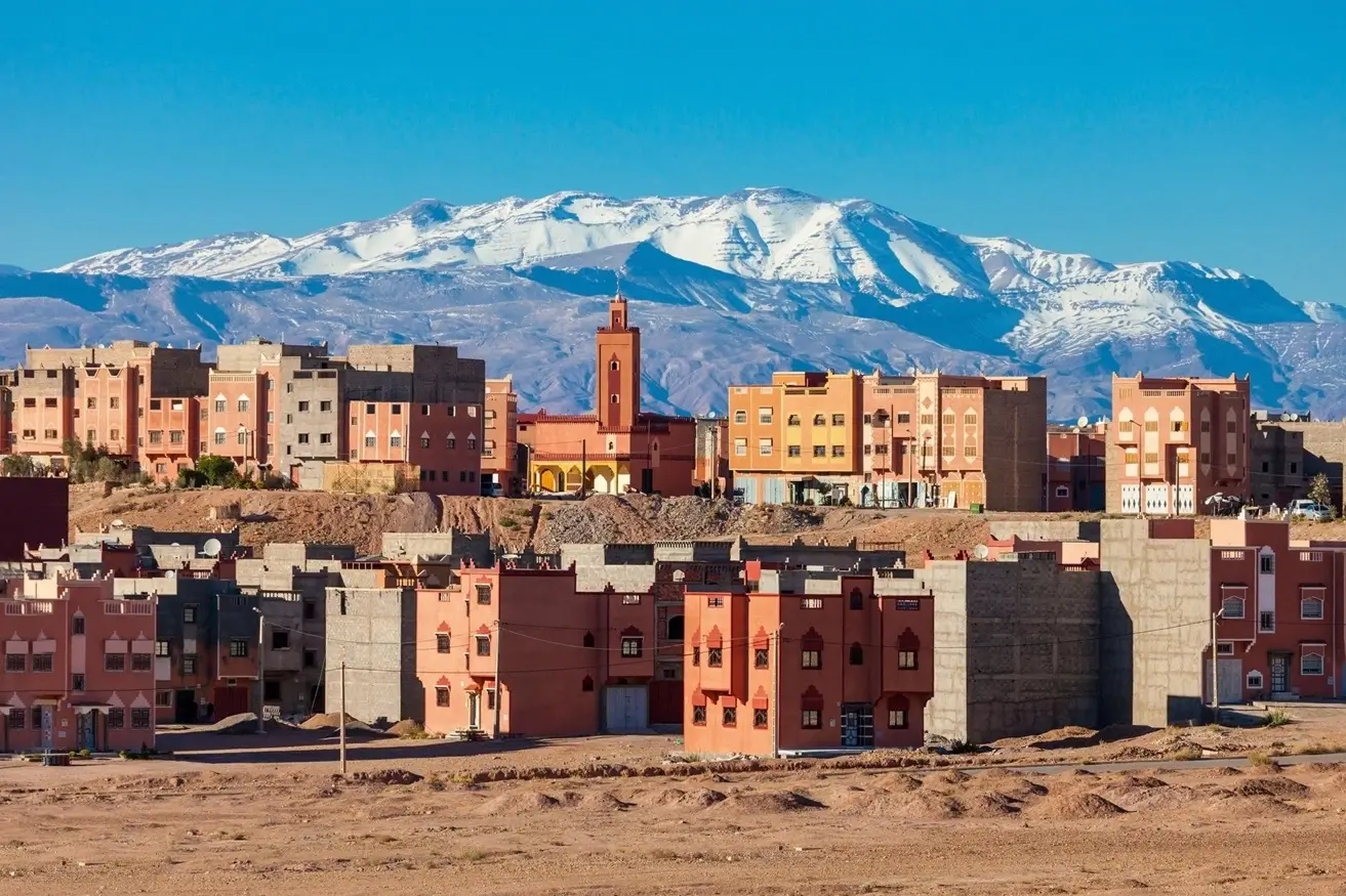 Scenic view of Ouarzazate, Morocco with the Atlas Mountains in the background. The image shows traditional Moroccan architecture with colorful buildings under a clear blue sky.