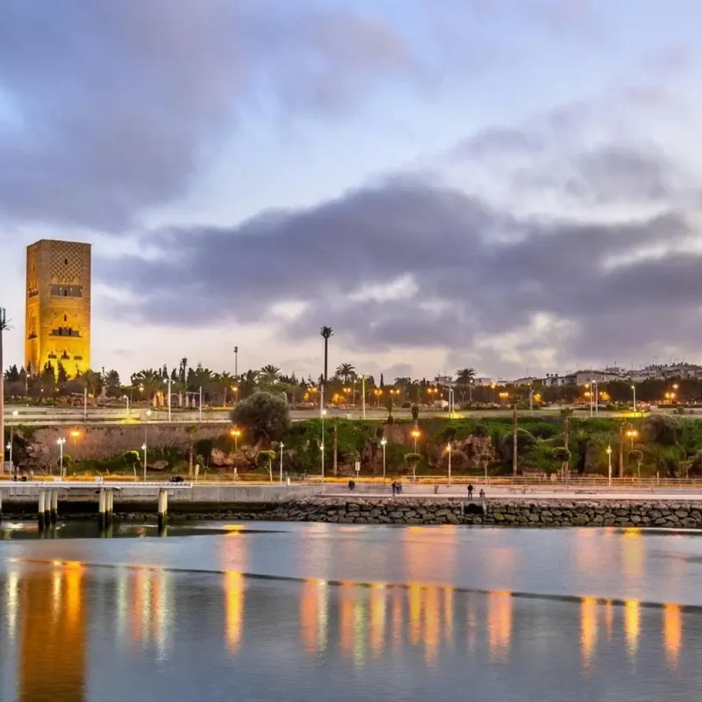 view of Rabat at dusk, with the Hassan Tower illuminated in the background and the reflections on the water adding to the charm of the city.