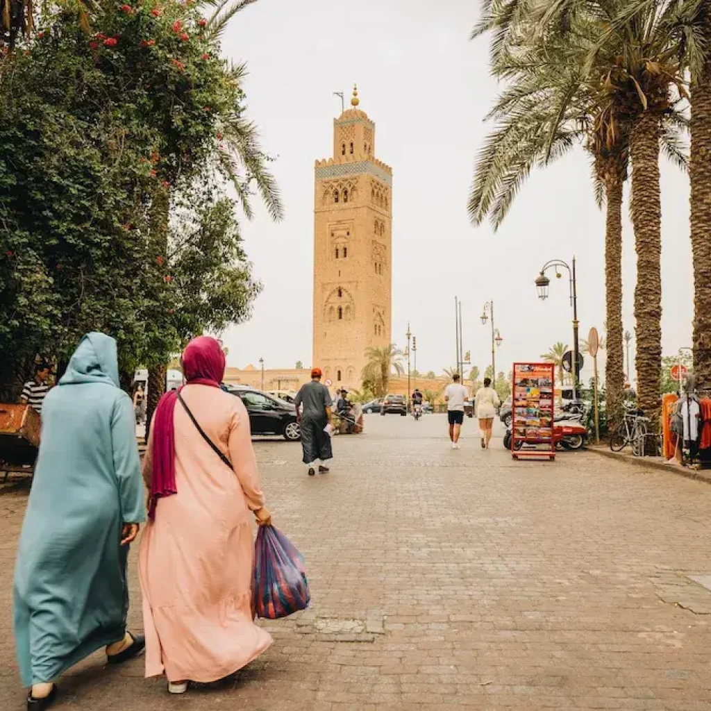 A street scene in Marrakech, Morocco, featuring two women in traditional Moroccan attire walking towards the Koutoubia Mosque. The tall minaret of the mosque stands prominently in the background. The street is lined with palm trees, shops, and vendors, contributing to the lively ambiance.