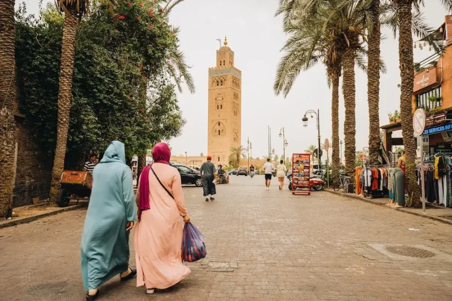 A street scene in Marrakech, Morocco, featuring two women in traditional Moroccan attire walking towards the Koutoubia Mosque. The tall minaret of the mosque stands prominently in the background. The street is lined with palm trees, shops, and vendors, contributing to the lively ambiance.