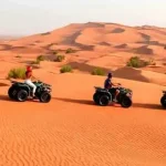 A group of seven people posing with their quad bikes on the sand dunes. They are dressed in protective gear and helmets, smiling and making victory signs with their hands. The background features sand dunes under a partly cloudy sky.