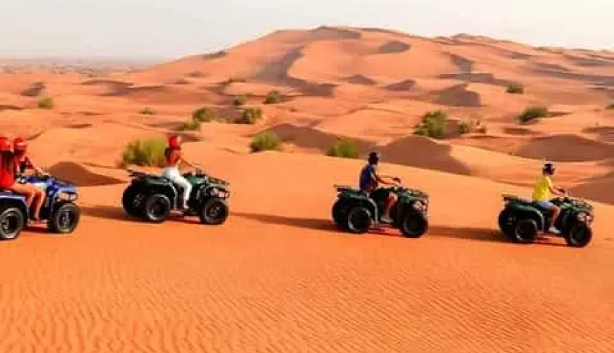 A group of seven people posing with their quad bikes on the sand dunes. They are dressed in protective gear and helmets, smiling and making victory signs with their hands. The background features sand dunes under a partly cloudy sky.