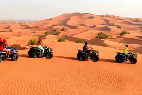 A group of seven people posing with their quad bikes on the sand dunes. They are dressed in protective gear and helmets, smiling and making victory signs with their hands. The background features sand dunes under a partly cloudy sky.