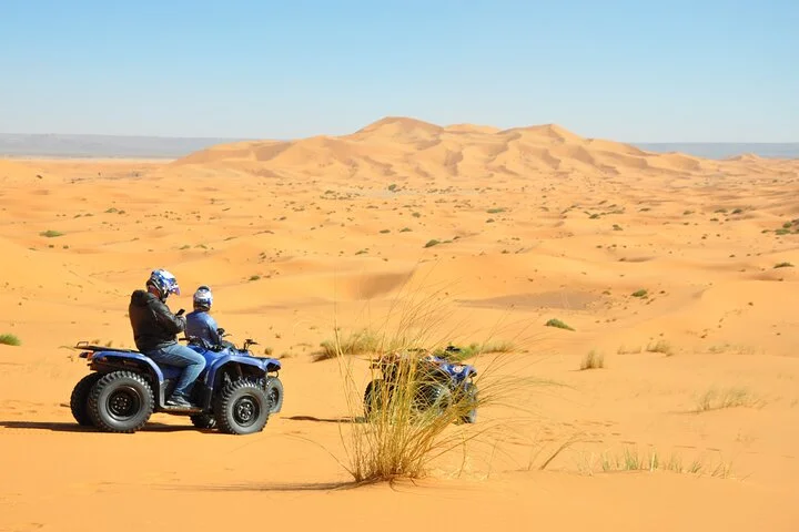 Two individuals on quad bikes pausing in a desert landscape, with expansive golden sand dunes stretching out under a clear blue sky.