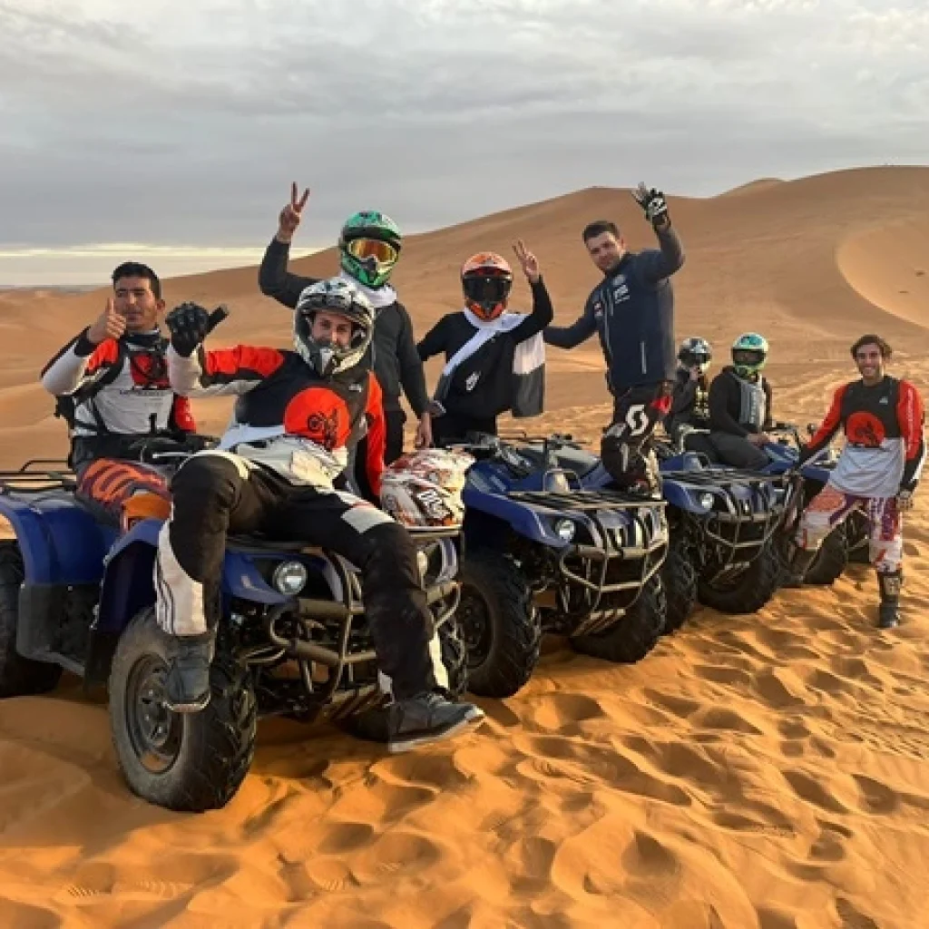 A group of people riding quad bikes in a single line formation across a vast desert landscape. The desert features rolling sand dunes, and the riders are wearing casual and protective clothing, including helmets.