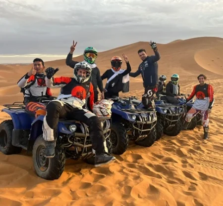 A group of people riding quad bikes in a single line formation across a vast desert landscape. The desert features rolling sand dunes, and the riders are wearing casual and protective clothing, including helmets.