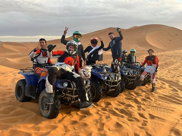 A group of people riding quad bikes in a single line formation across a vast desert landscape. The desert features rolling sand dunes, and the riders are wearing casual and protective clothing, including helmets.