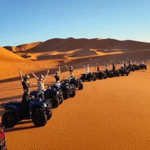 Un grupo de personas montan en quads en fila india por un vasto desierto de arena con dunas ondulantes al fondo.