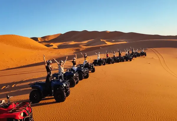 A group of people riding quad bikes in a single file across a vast, sandy desert with rolling dunes in the background.