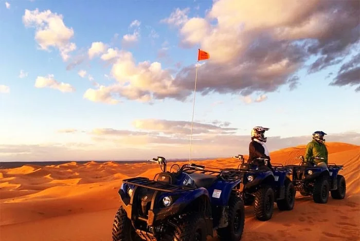 Two individuals on quad bikes, paused on the edge of a sand dune in a desert setting. They are wearing helmets and protective gear, with the vast expanse of sand dunes stretching out behind them under a clear blue sky.