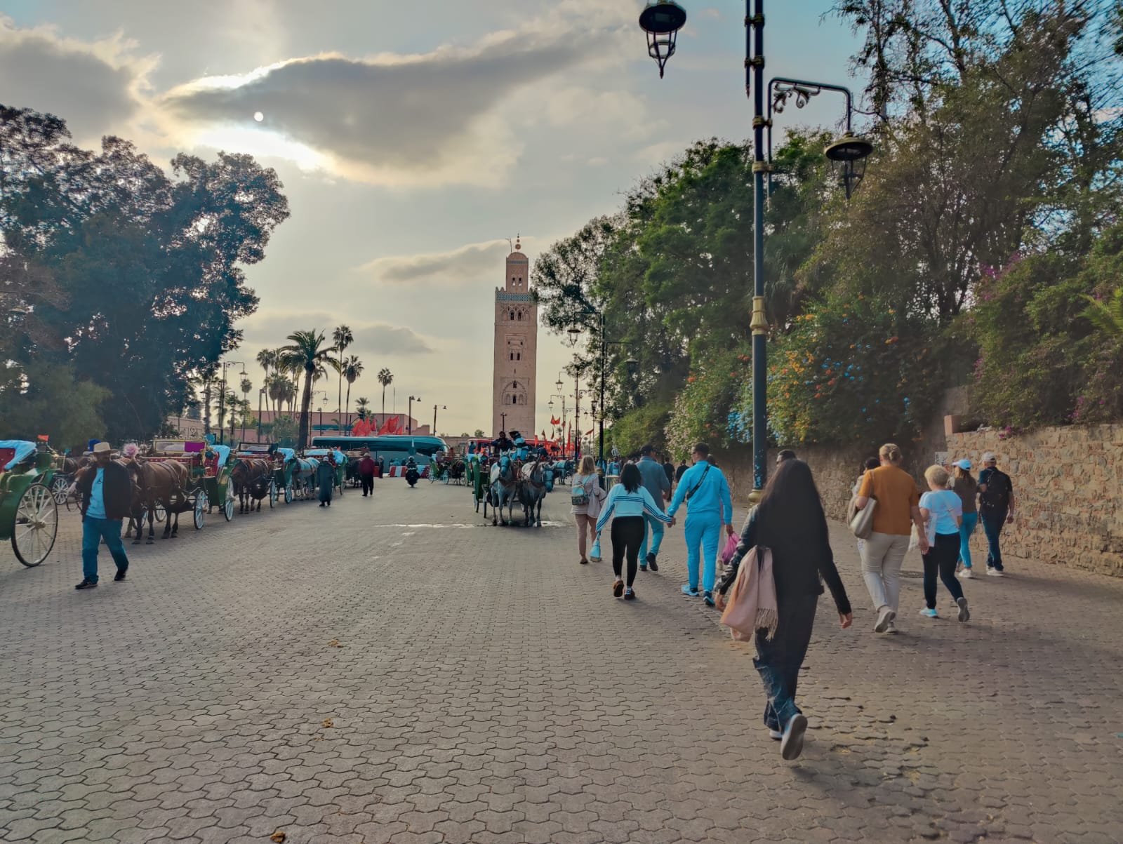 Calle concurrida cerca de la mezquita de Koutoubia en Marrakech, Marruecos, con turistas paseando y coches de caballos esperando. El cielo está parcialmente nublado y los árboles con flores de colores bordean el camino. Un lugar perfecto para hacer turismo por Marrakech y explorar la cultura marroquí.