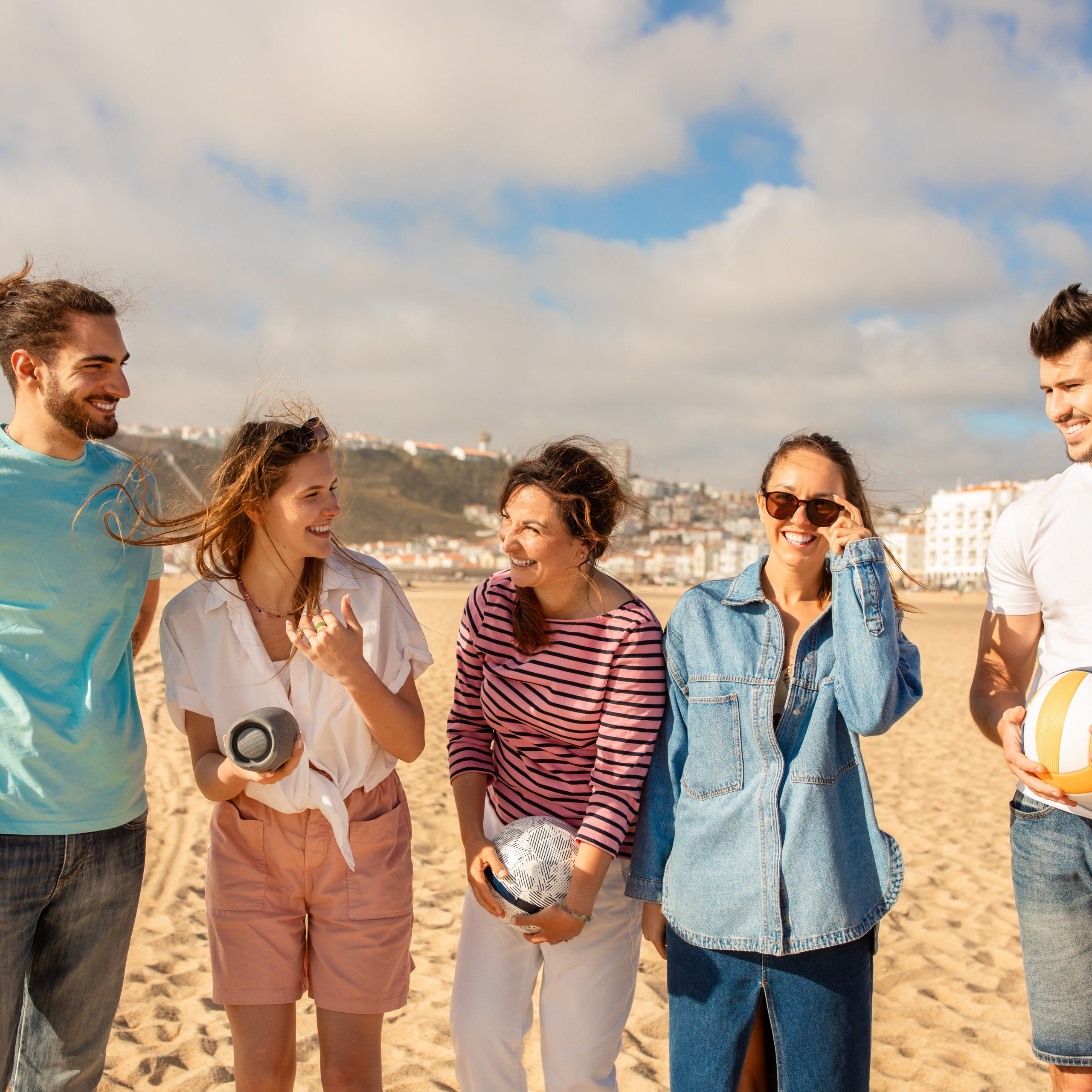 A group of five friends enjoys a lively day at the beach, engaging in playful activities and sharing laughter against a backdrop of clouds and coastline scenery.