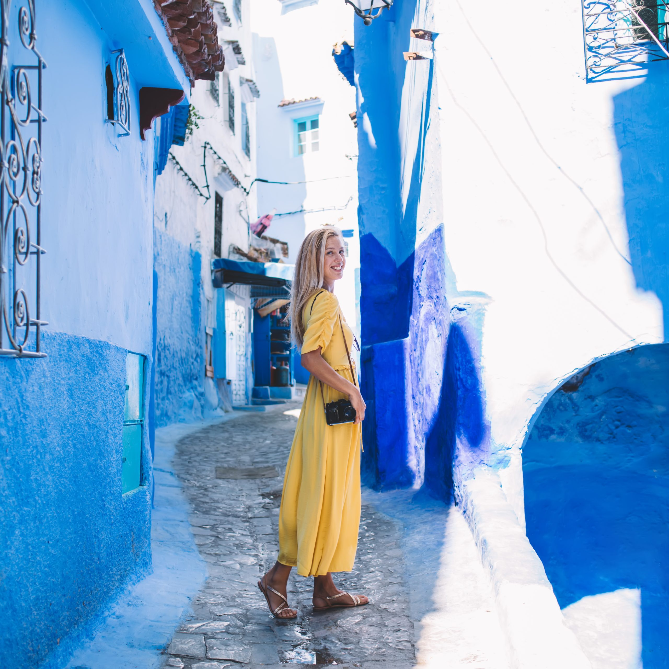 Full body glad female photographer smiling and looking at camera while standing on stone path outside shabby houses on sunny day in Morocco
