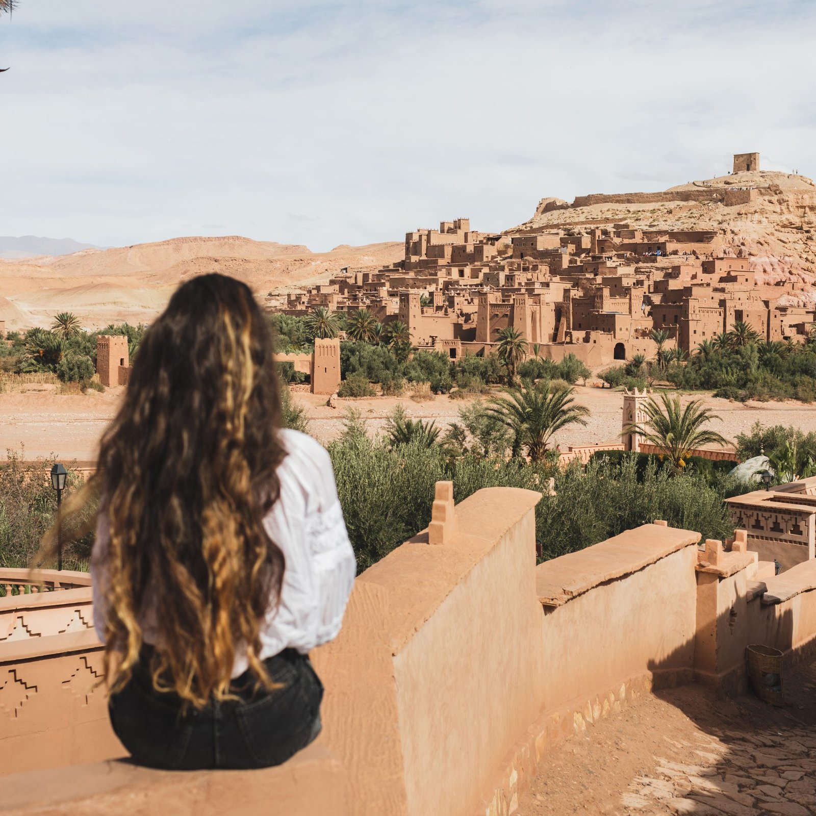 Woman sitting on fence with view of famous moroccan old town ksar Ait-Ben-Haddou. View from behind. Welcome to Morocco, Ouarzazate.