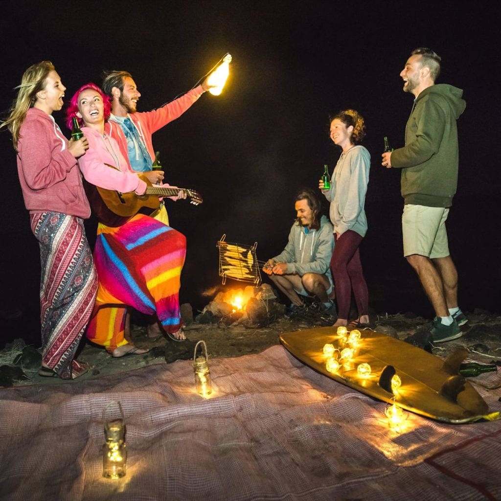 Young hipster friends having fun together at beach camping party with night campfire light - Friendship travel concept with young surfer people grilling corn cob at summer barbecue